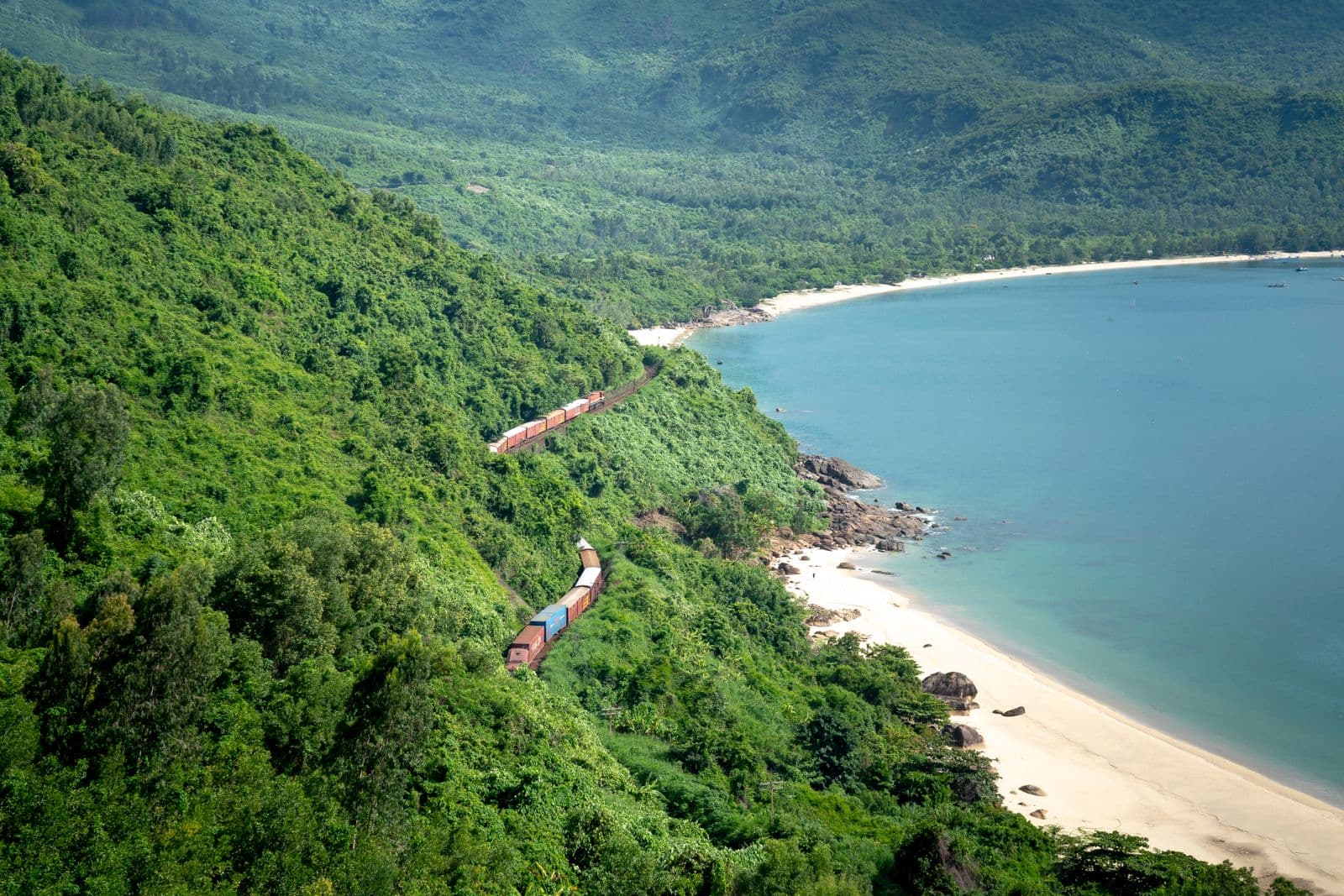 A train winding along the coastal railway over the Hai Van Pass, between forested mountains and a turquoise bay near Da Nang, Vietnam.